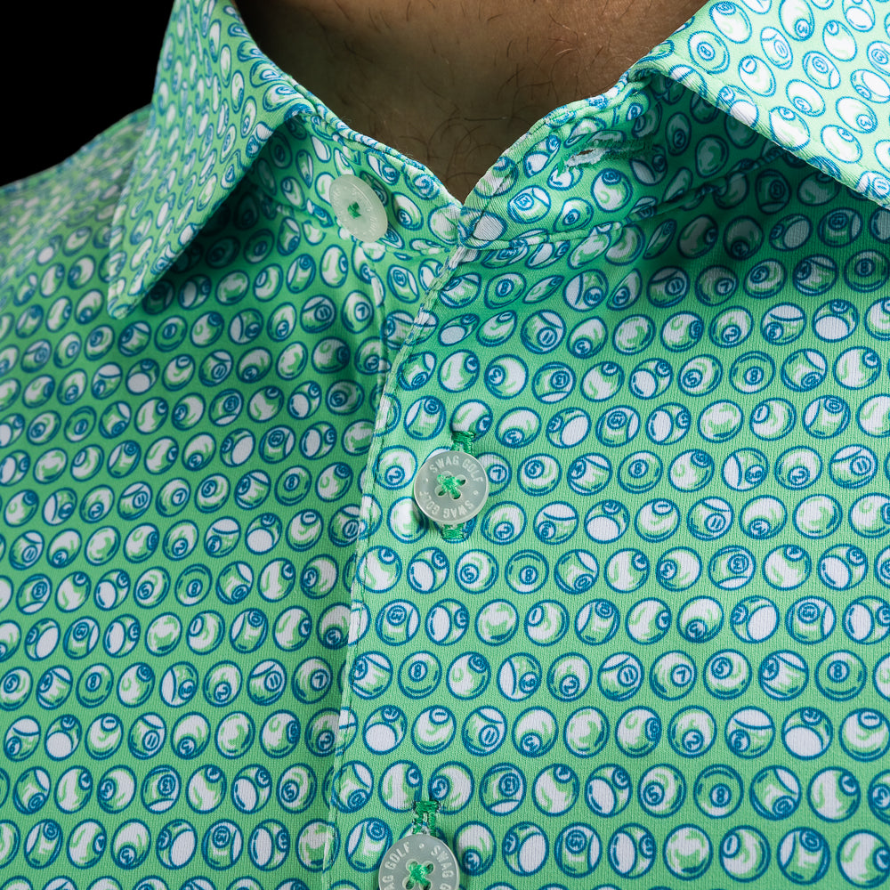 Close-up of a men's golf polo shirt with a green base and a repeating blue and white 8-ball pool pattern, worn by a model.