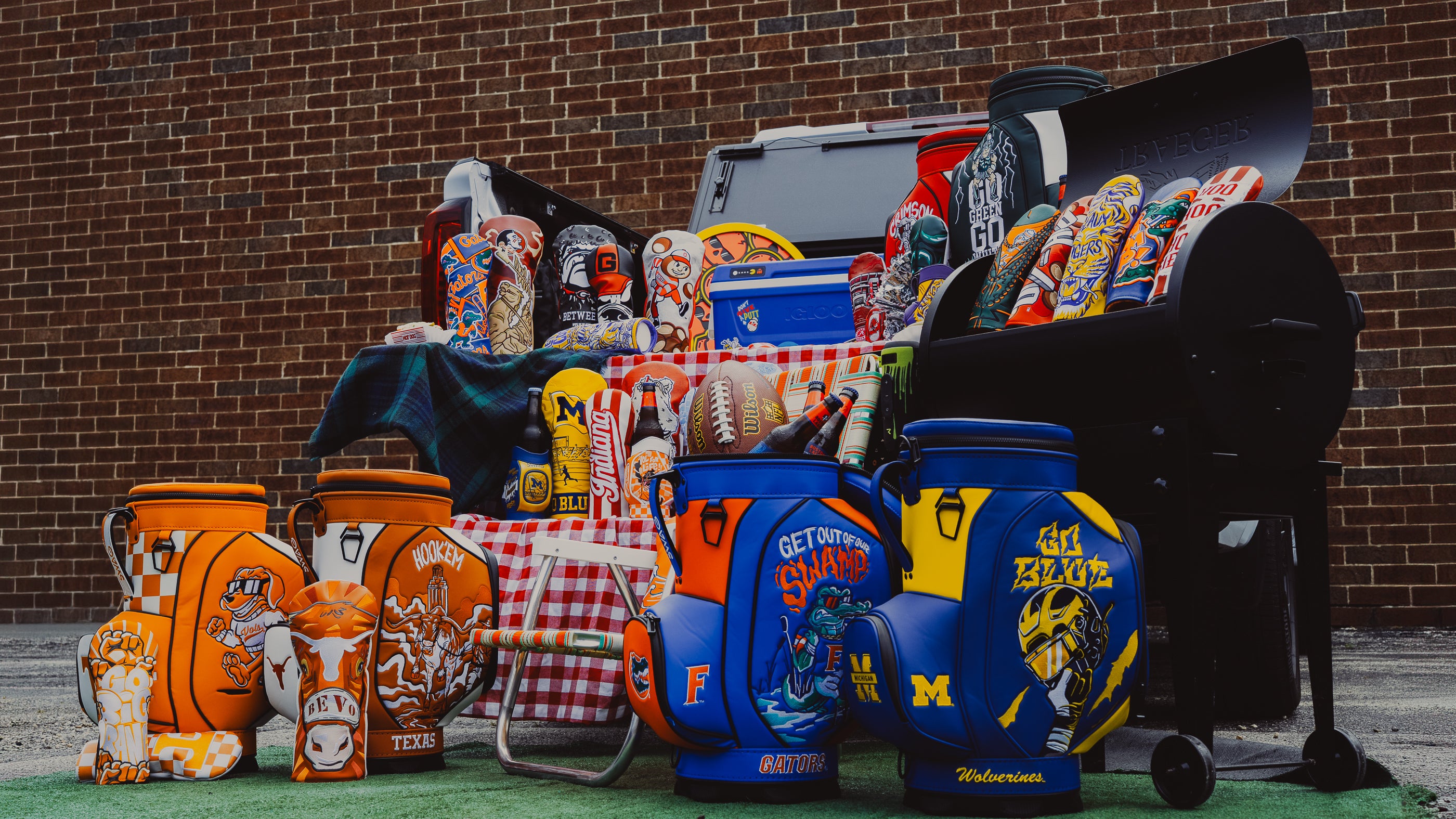 A collection of sports-themed golf headcovers, desk caddies and cooler bags displayed outdoors in front of a brick wall. The items feature various collegiate logos, including Texas, Michigan State, and Florida, along with red-and-white checkered tablecloths and a black grill in the background.