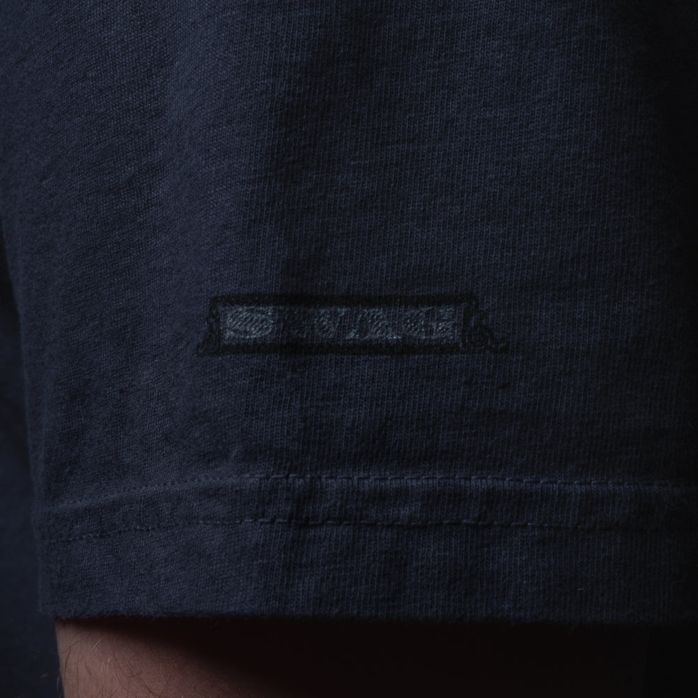 Close-up of tonal black script SWAG logo printed on sleeve of navy Blackout Bills Jefferson t-shirt.