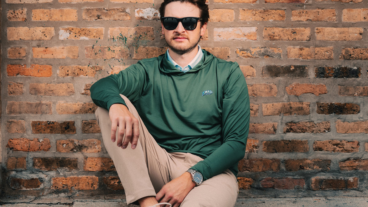 Model wearing olive green long-sleeve golf hoodie sitting against a brick wall.