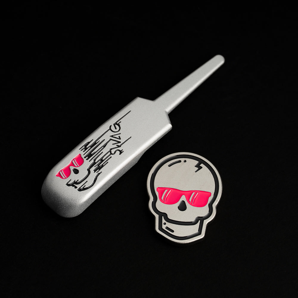 Silver metal SWAG divot repair tool engraved with skull logo and matching magnetic skull ball marker featuring bright pink sunglasses, photographed on a black background.