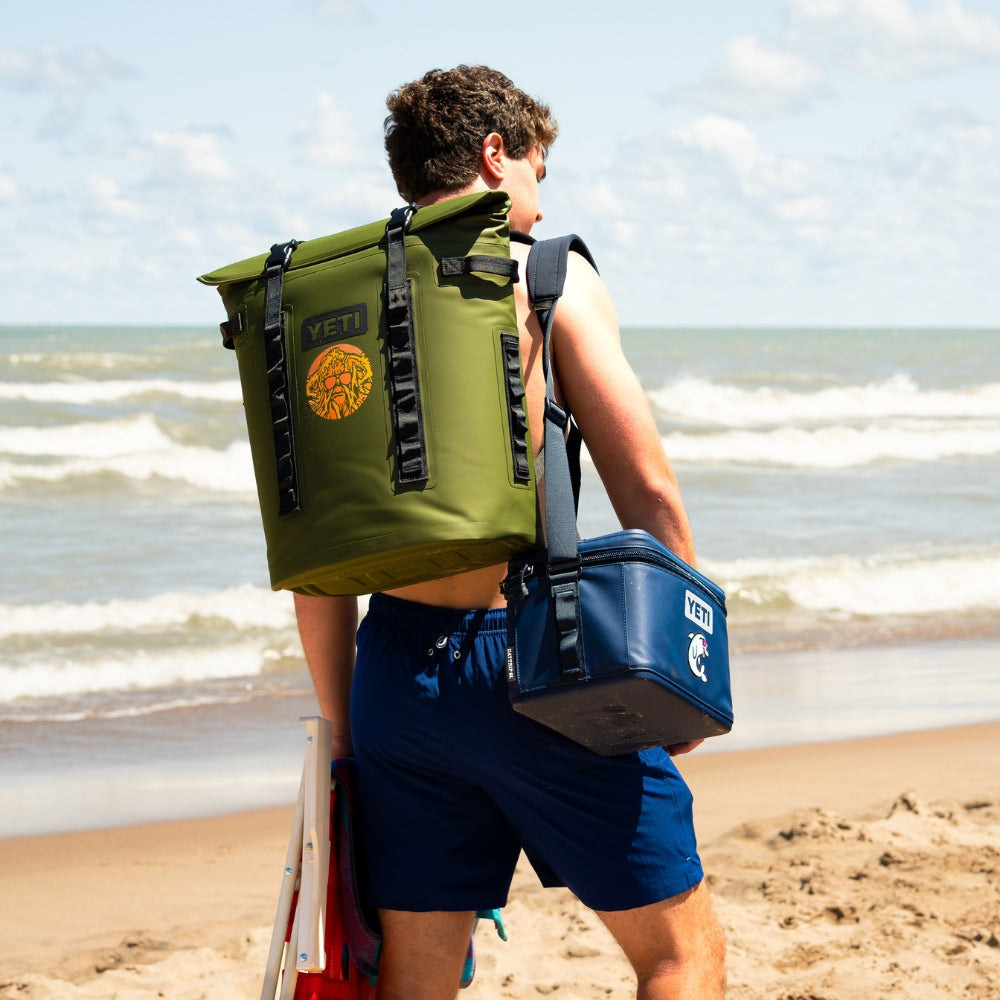 A man on a sandy beach wearing a large olive green YETI backpack and carrying a blue YETI cooler bag over his shoulder, with ocean waves in the background.