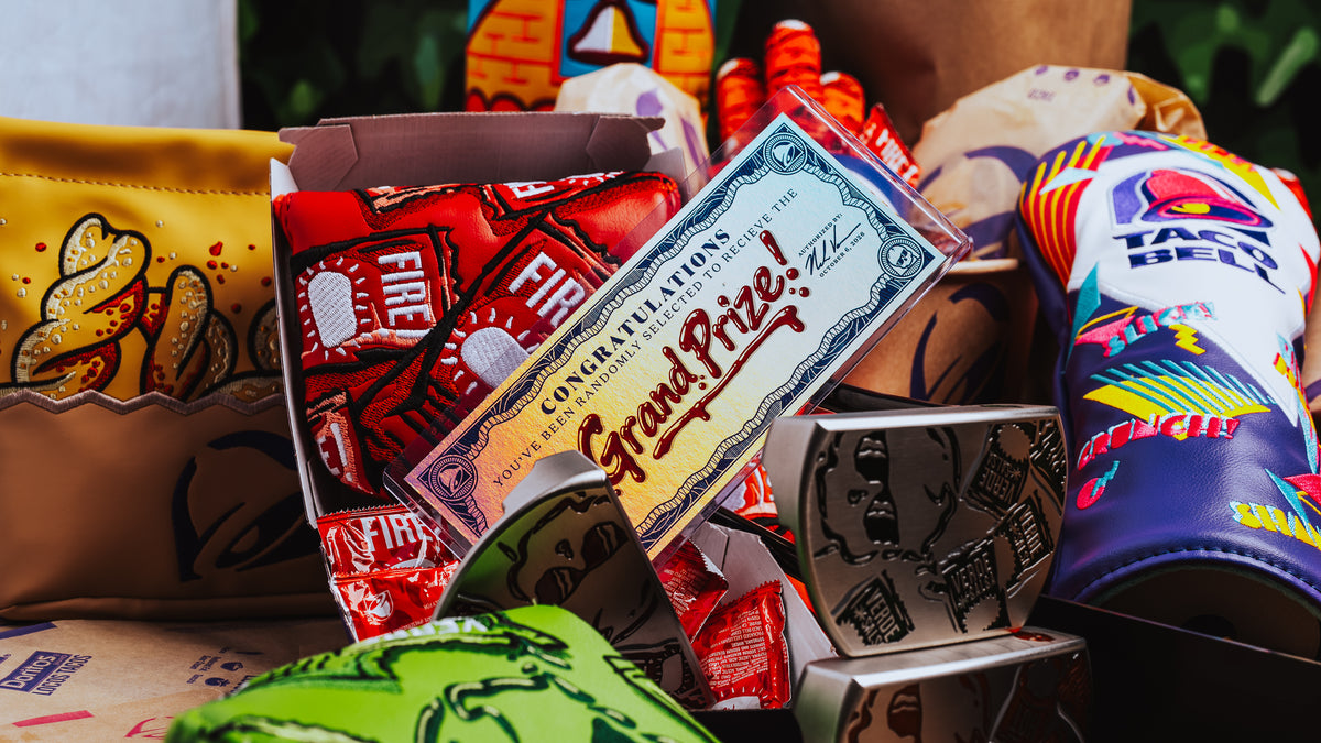 A close-up, styled flat-lay of various novelty pouches and small boxes, featuring a central cream-colored 'Grand Prize' certificate and surrounding colorful items including a yellow patterned pouch, a red 'Fire' pouch, and a blue 'Taco Bell' pouch.