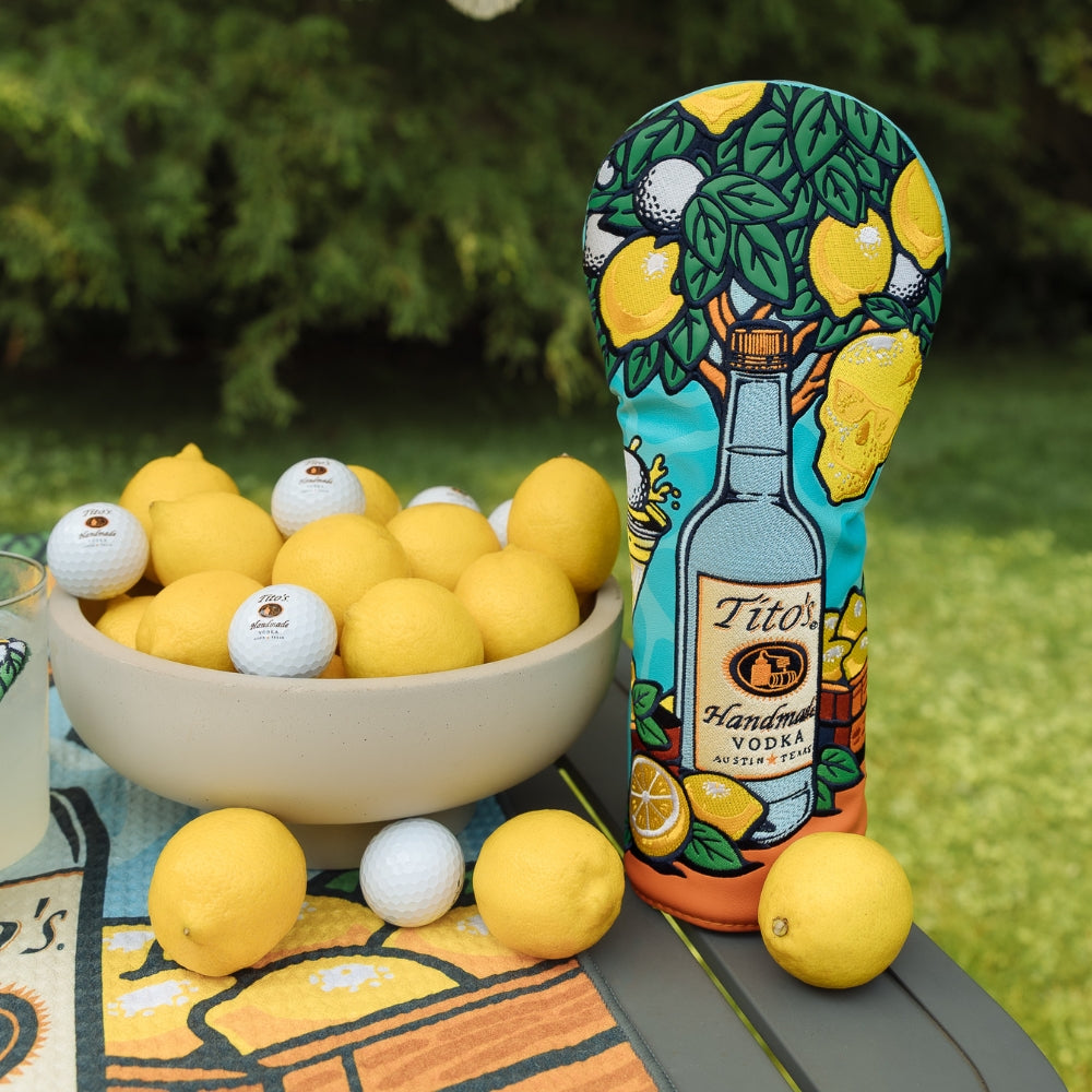 A golf club headcover featuring a bottle of Tito's Handmade Vodka stands on a table outdoors, surrounded by lemons and golf balls. The headcover features a colorful illustration of a lemon tree and the bottle graphic.