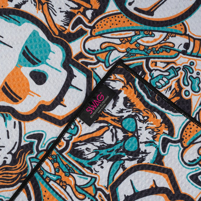 White SWAG golf towel with full-coverage teal and orange skull and king artwork in a bold graphic collage pattern.