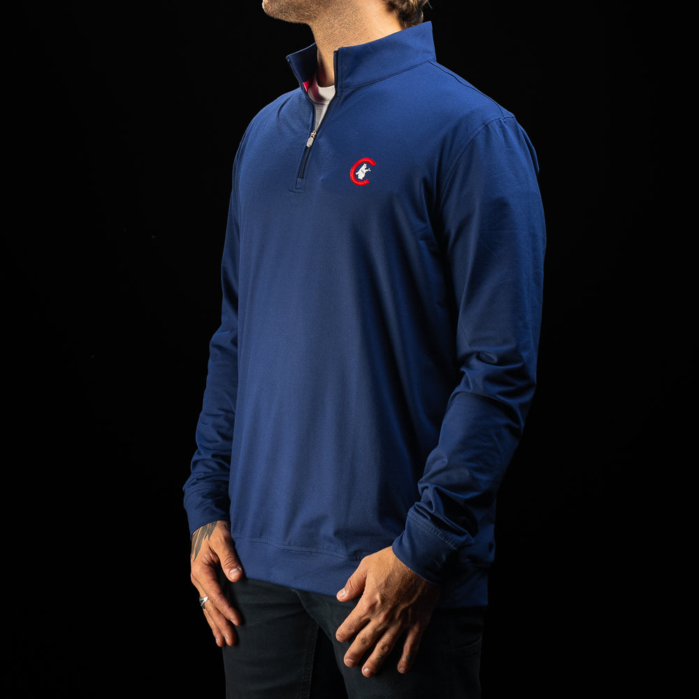 A man wearing a royal blue quarter-zip pullover sweater with a small red and white embroidered logo on the left chest, shown against a solid black background.