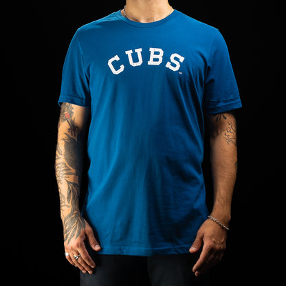 A man wearing a bright blue short-sleeve t-shirt with 'CUBS' printed in white arched block letters across the chest, shown against a solid black background.