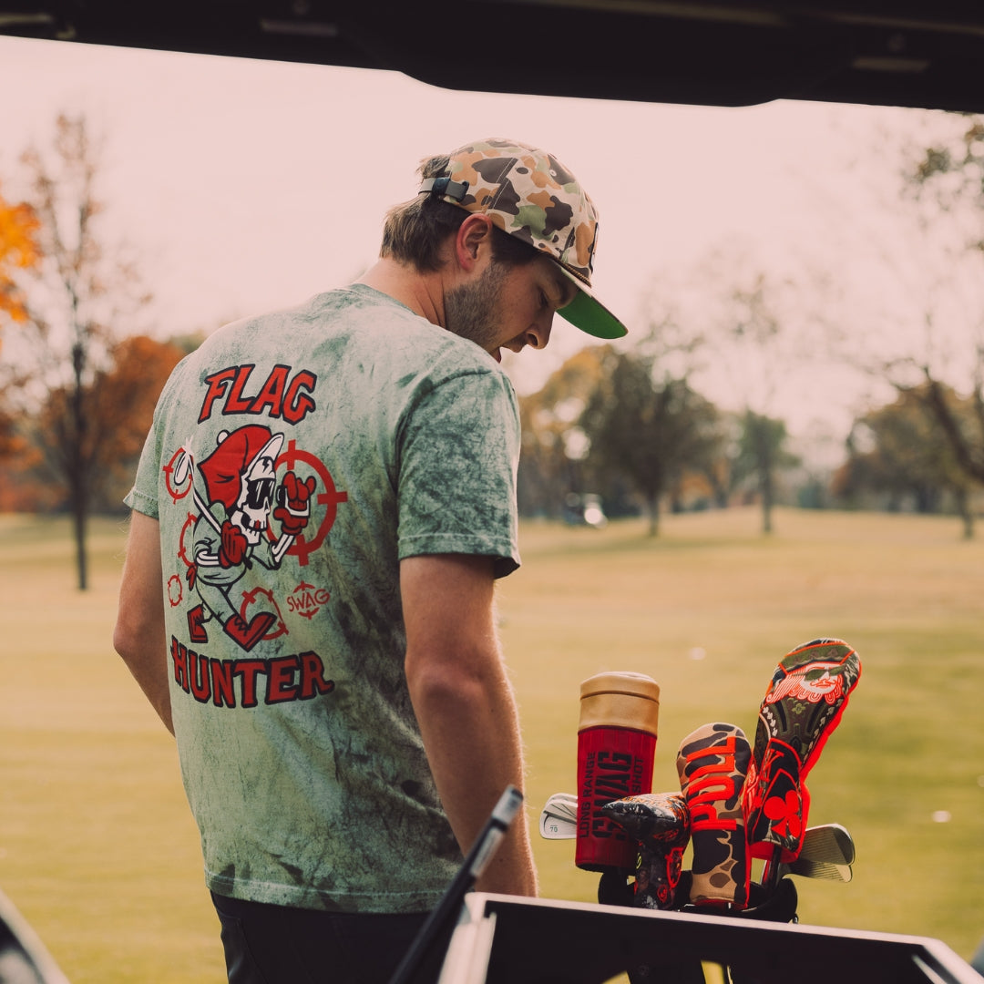 SWAG Golf model wearing a green and white camouflage Flag Hunter garment dyed men's short sleeve golf T-Shirt with orange target and white flag pin on the left chest. 