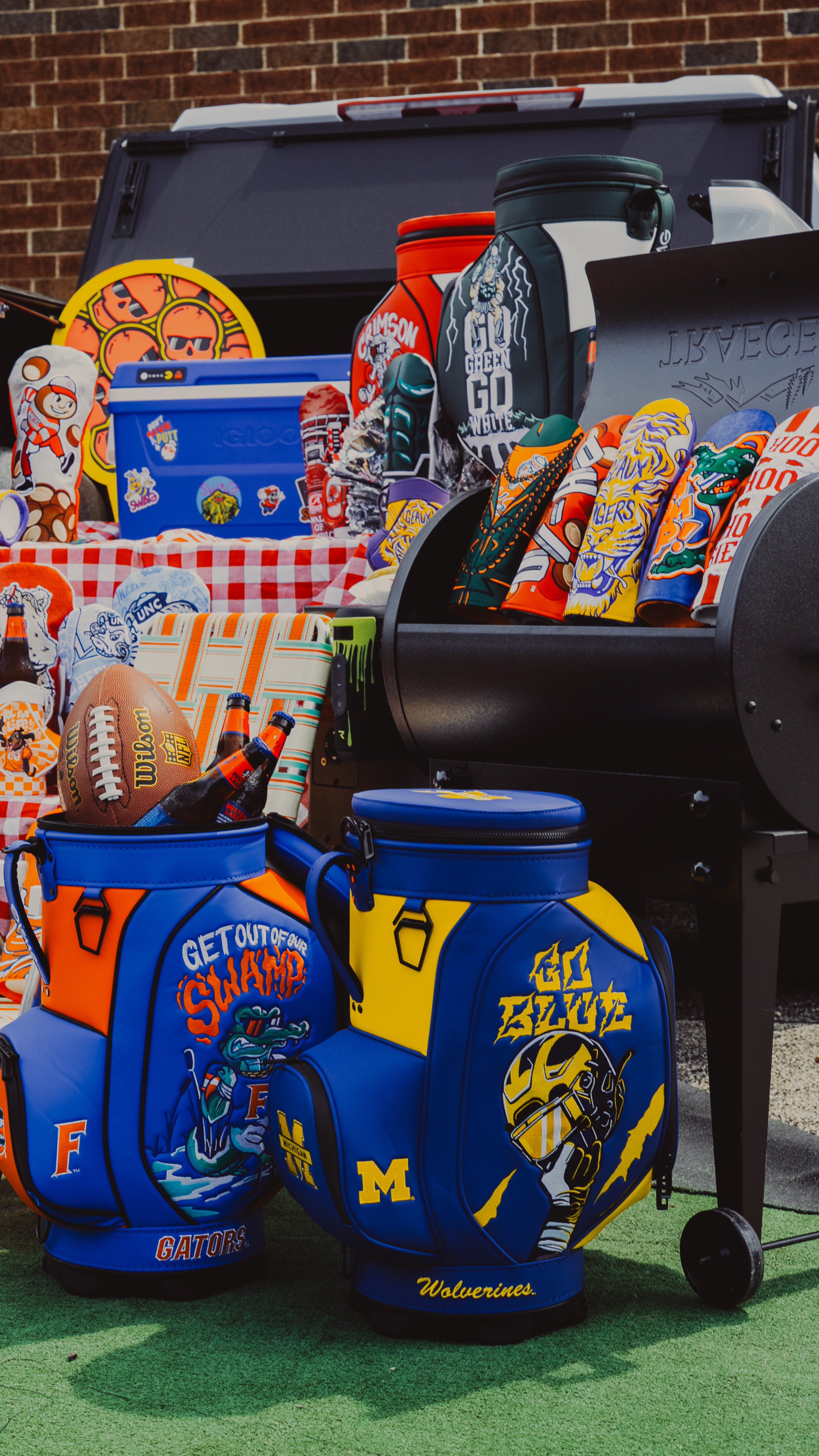 A collection of sports-themed golf headcovers, desk caddies and cooler bags displayed outdoors in front of a brick wall. The items feature various collegiate logos, including Texas, Michigan State, and Florida, along with red-and-white checkered tablecloths and a black grill in the background.