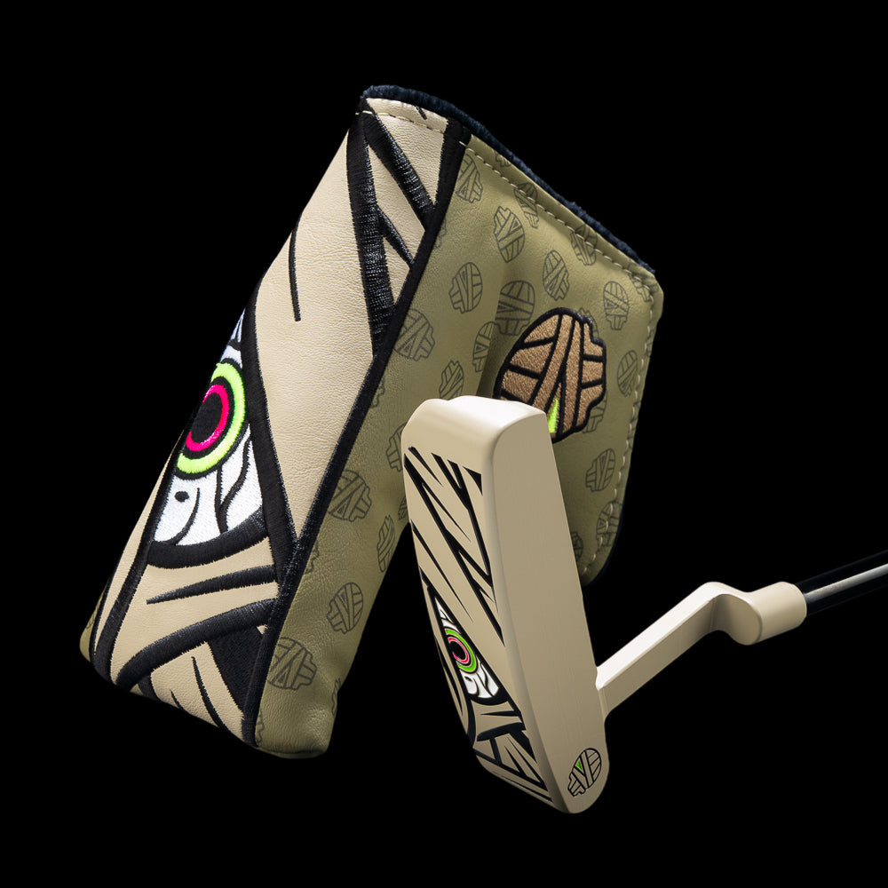 A Halloween Mummy themed golf putter and its matching cover set against a solid black background. The putter is cream-colored with black graphic stripes and a green and pink eye design, while the cover features a matching beige and black patterned design.