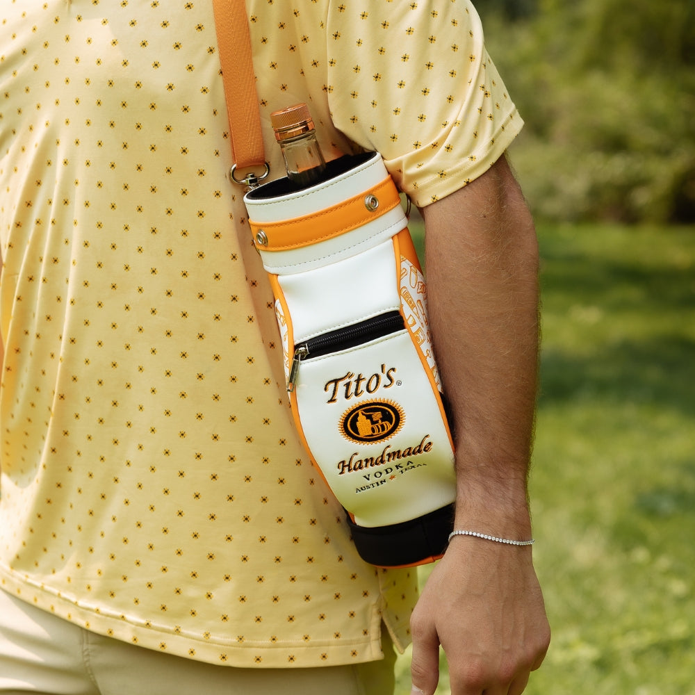 A white and orange Tito's bottle carrier sling bag with a Tito's Handmade logo, worn crossbody on a person wearing a yellow patterned polo shirt in an outdoor grassy setting.