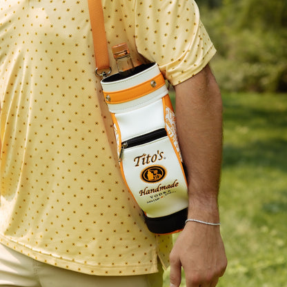 A white and orange Tito's bottle carrier sling bag with a Tito's Handmade logo, worn crossbody on a person wearing a yellow patterned polo shirt in an outdoor grassy setting.