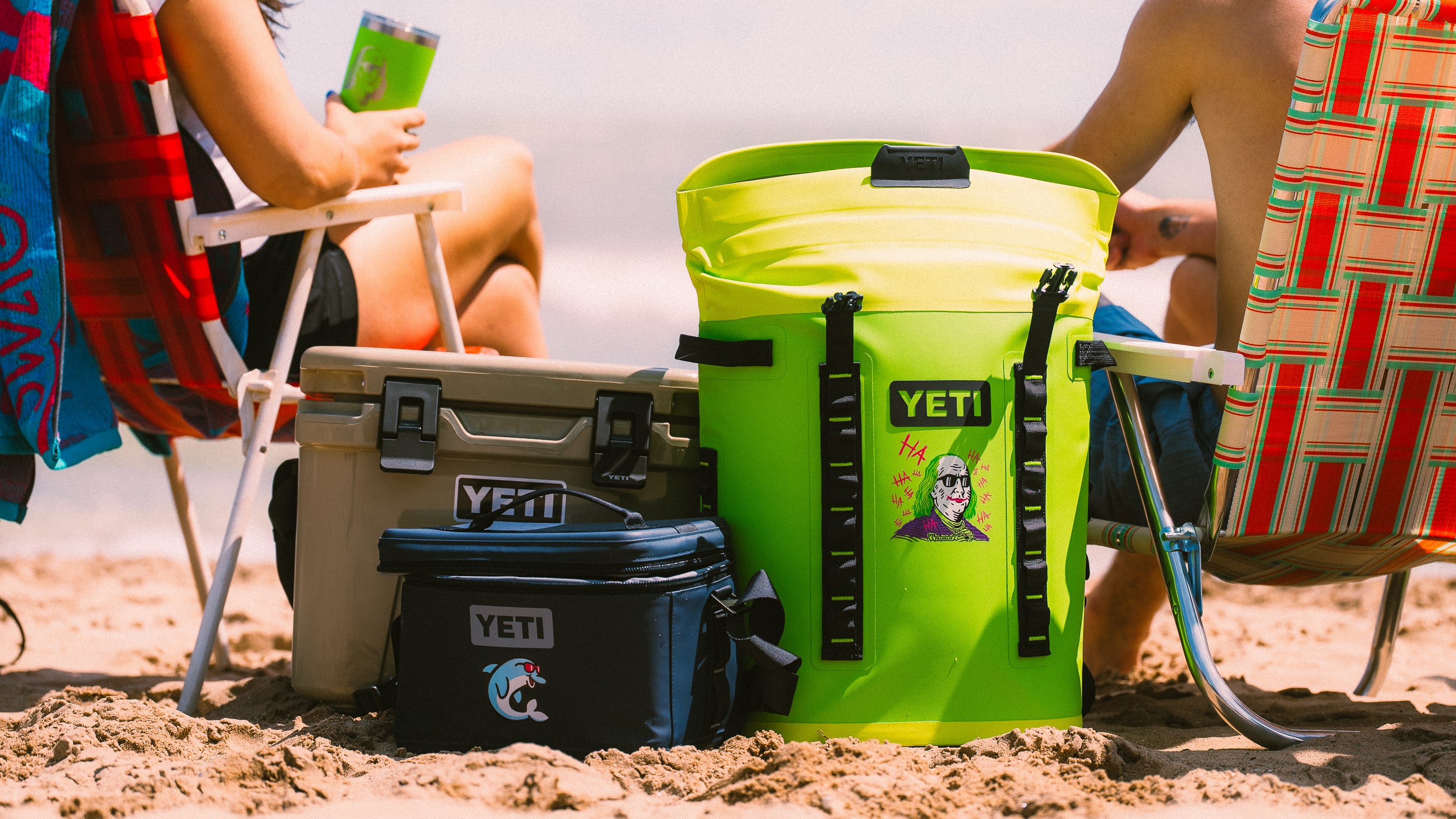 A bright lime green YETI cooler bucket with black straps and a cartoon sticker, standing on sand next to a gray YETI cooler and a dark blue YETI hard cooler, with beachgoers seated in folding chairs at a beach.